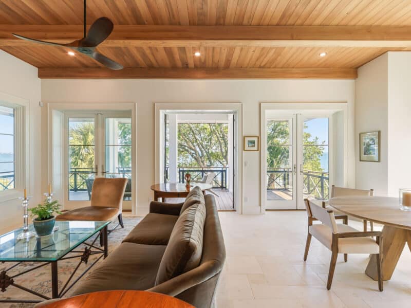 An open-concept living and dining room featuring a wood-plank ceiling, travertine floors, and large French doors opening to a scenic waterfront treescape deck.
