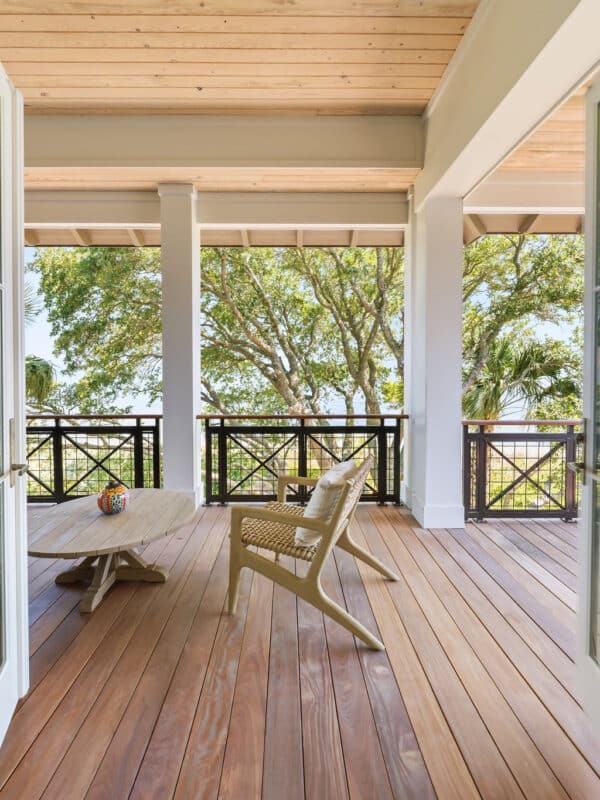 A spacious covered wooden porch with a light-colored wood floor, white support pillars, and a light wood plank ceiling. The porch features two modern woven armchairs, a low oval wooden coffee table, and a dark metal railing looking out at trees that frame a waterfront view.
