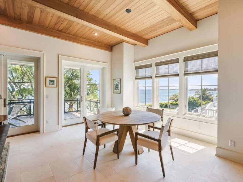 Bright dining area featuring a round wooden table with four white upholstered chairs, tile flooring, a natural wood-planked ceiling with exposed beams, and large windows offering a scenic view of a coastal bay.