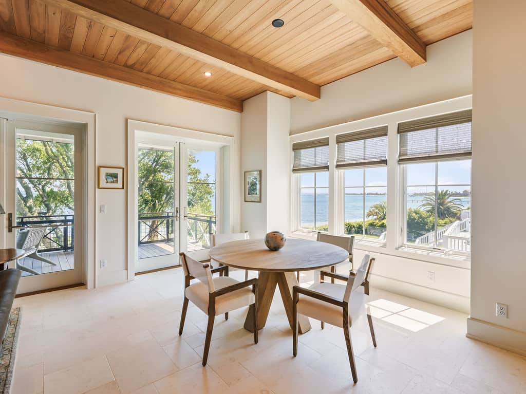 Bright dining area featuring a round wooden table with four white upholstered chairs, tile flooring, a natural wood-planked ceiling with exposed beams, and large windows offering a scenic view of a coastal bay.