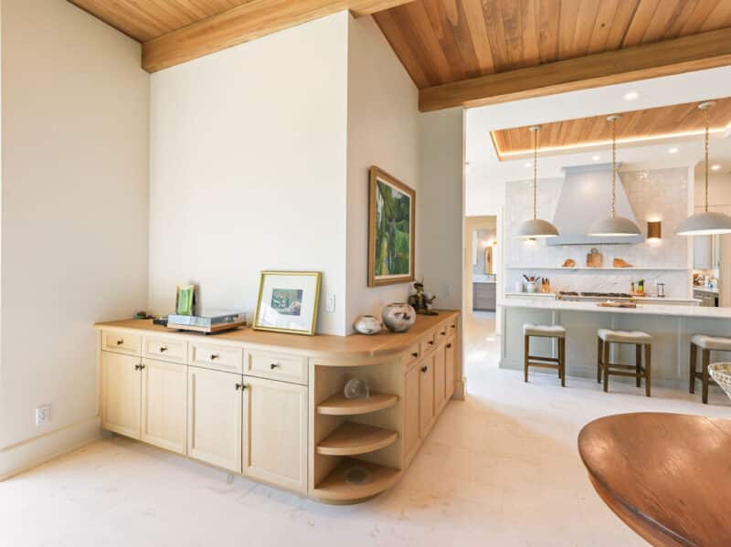 Light wood buffet cabinet wrapping around a white wall in an open-concept home next to a kitchen with a large island and wood-beamed ceilings.
