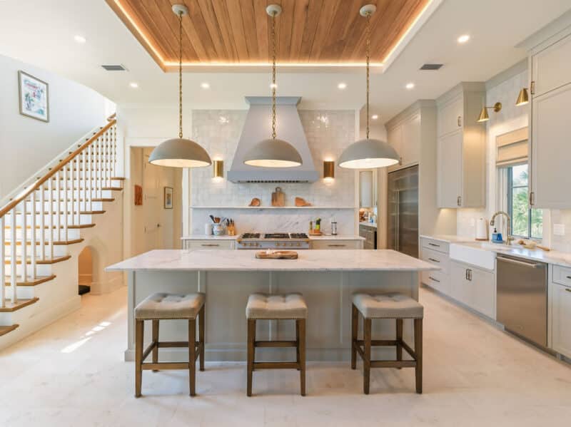 Modern luxury kitchen featuring a large marble island with three barstools, a light blue range hood, a wooden tray ceiling with pendant lights, and an adjacent wooden staircase.
