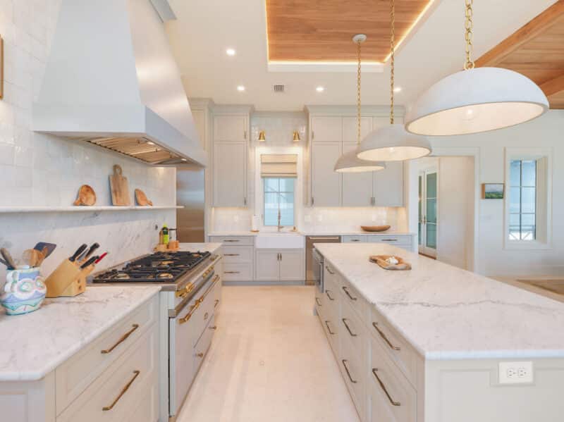 Modern luxury kitchen featuring a large marble island, three dome pendant lights, light gray cabinetry, a gas range, and a wood-accented ceiling.