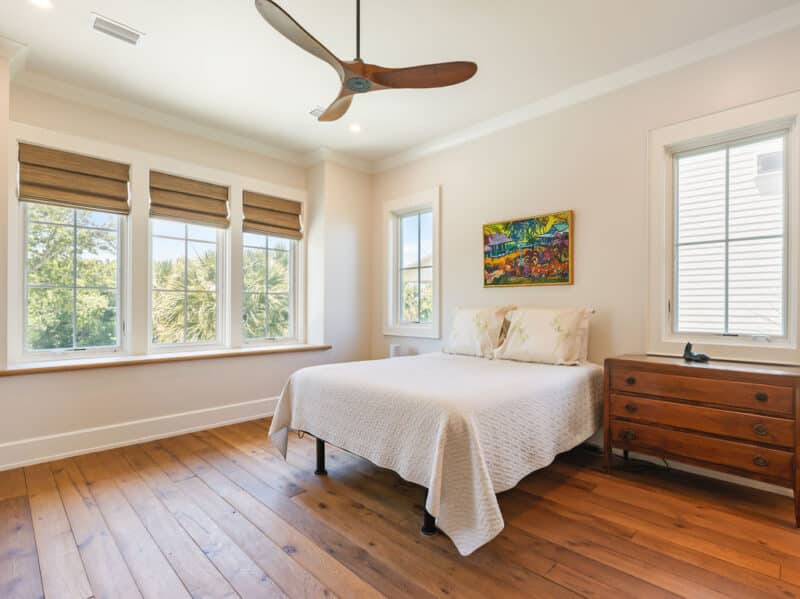 A bright, airy bedroom featuring light-colored walls, wide-plank hardwood floors, a white quilted bed, a wooden three-blade ceiling fan, and large multi-pane windows.