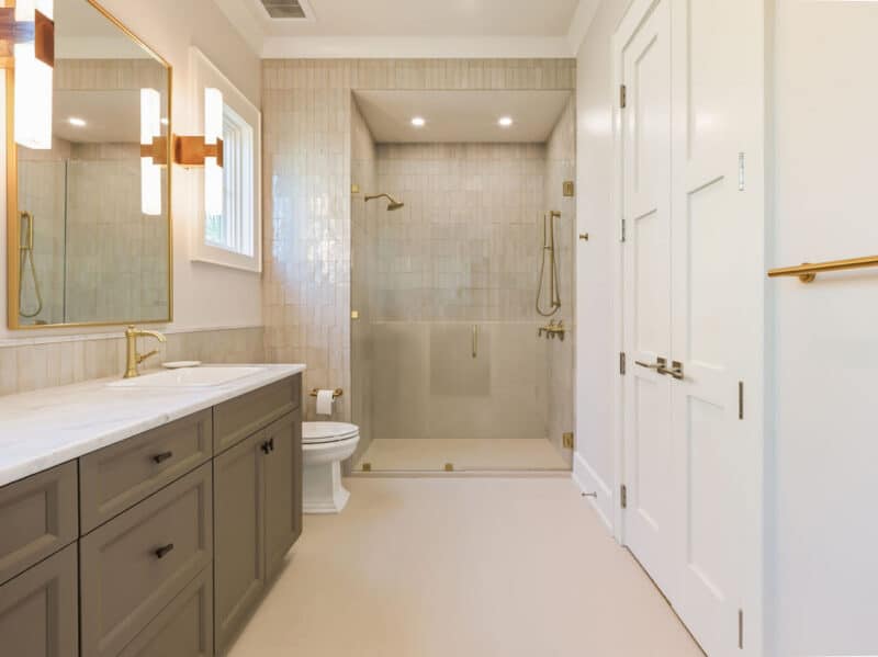 Modern primary bathroom featuring a taupe double vanity with a white marble countertop, gold hardware, a large walk-in glass shower with beige subway tiles, and white interior double doors.