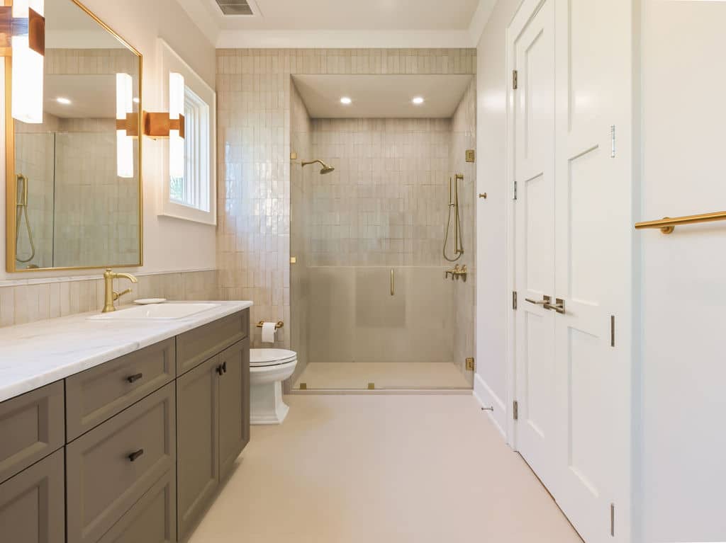 Modern primary bathroom featuring a taupe double vanity with a white marble countertop, gold hardware, a large walk-in glass shower with beige subway tiles, and white interior double doors.