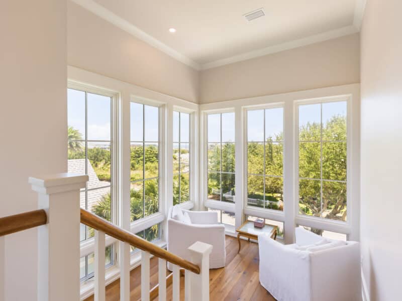 Bright sunlit seating nook at the top of a staircase featuring two white club chairs, hardwood floors, and large multi-pane windows overlooking lush coastal greenery.