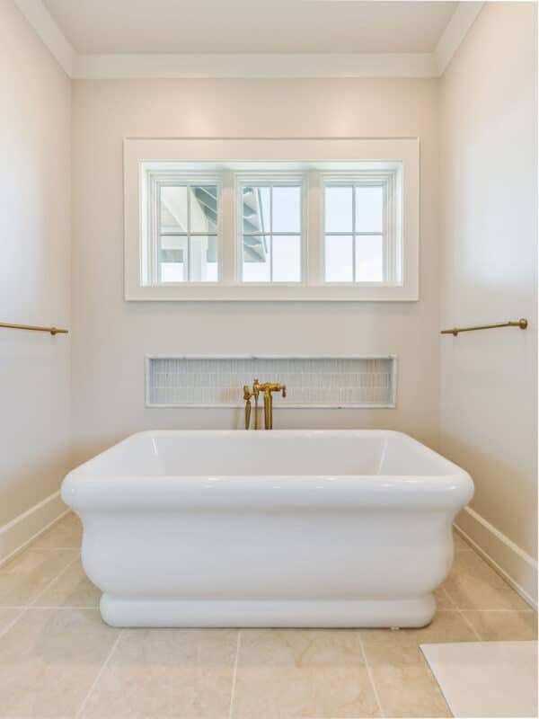 A modern white freestanding soaking tub with a floor-mounted brushed gold faucet, positioned beneath a three-pane window in an intimate bathroom nook.