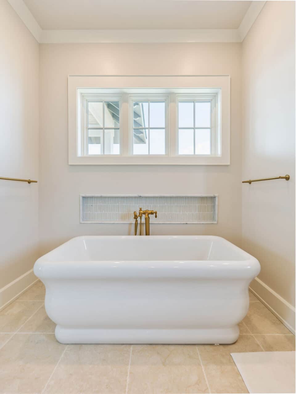 A modern white freestanding soaking tub with a floor-mounted brushed gold faucet, positioned beneath a three-pane window in an intimate bathroom nook.