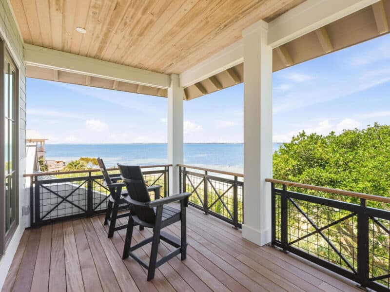 Two grey outdoor chairs on a spacious wooden covered balcony with a black railing, overlooking an ocean bay.