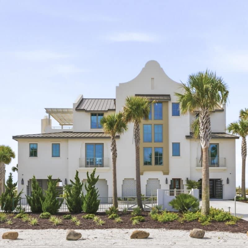 A modern three-story white luxury beach house with palm trees and scalloped parapet roofline..