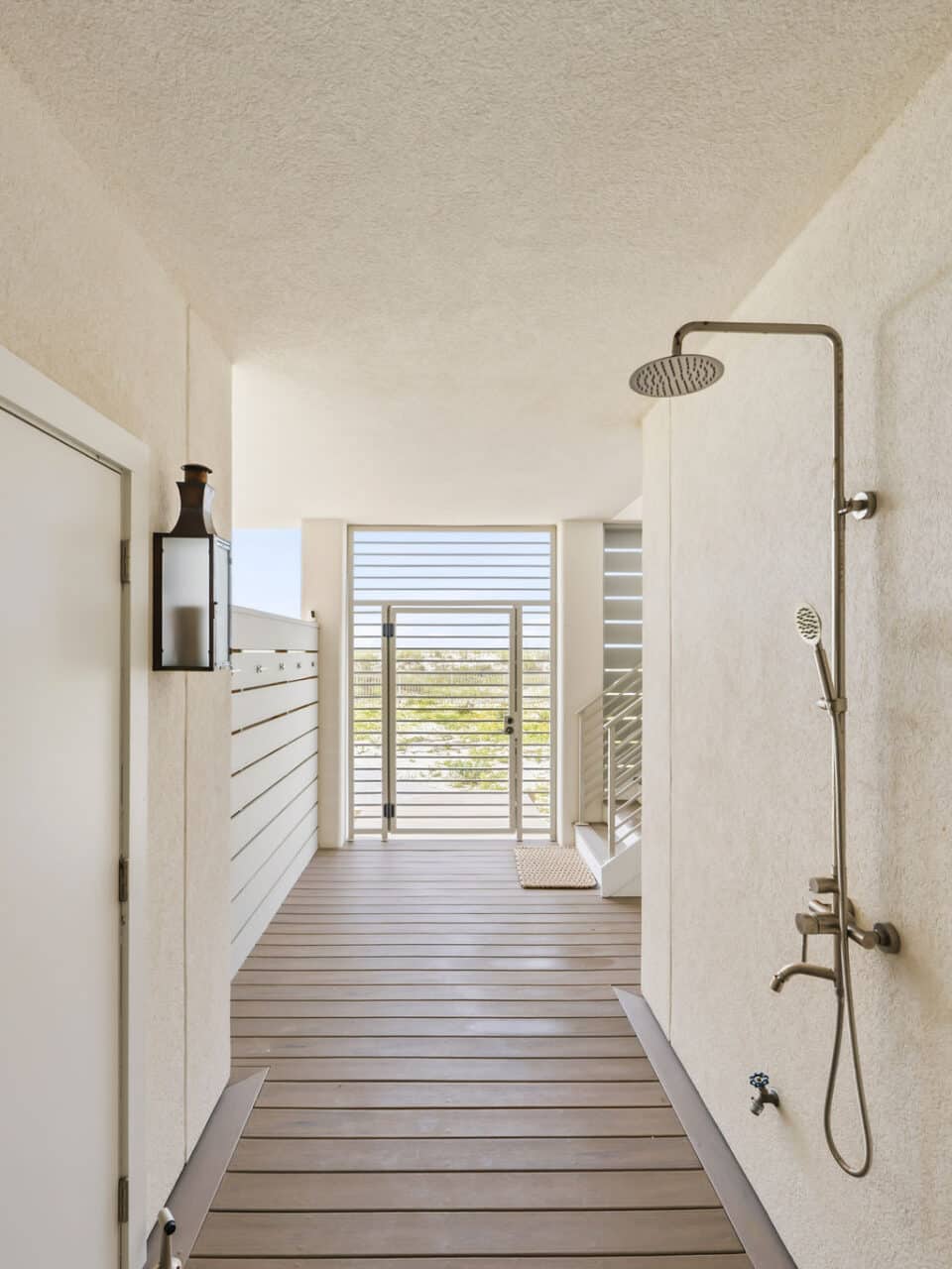 Modern outdoor, covered walkway with a shower and stainless steel rainfall showerhead, leading to a slatted metal beach gate.