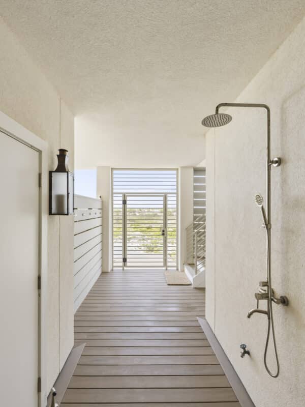 Modern outdoor, covered walkway with a shower and stainless steel rainfall showerhead, leading to a slatted metal beach gate.