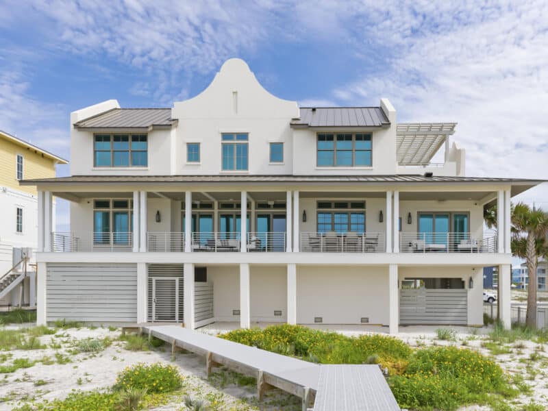 Three-story white beach house with a Dutch colonial-style roofline, a wide second-floor covered balcony, and a boardwalk to the beach.