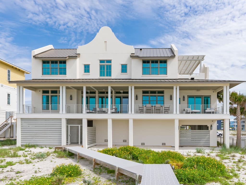 Three-story white beach house with a Dutch colonial-style roofline, a wide second-floor covered balcony, and a boardwalk to the beach.