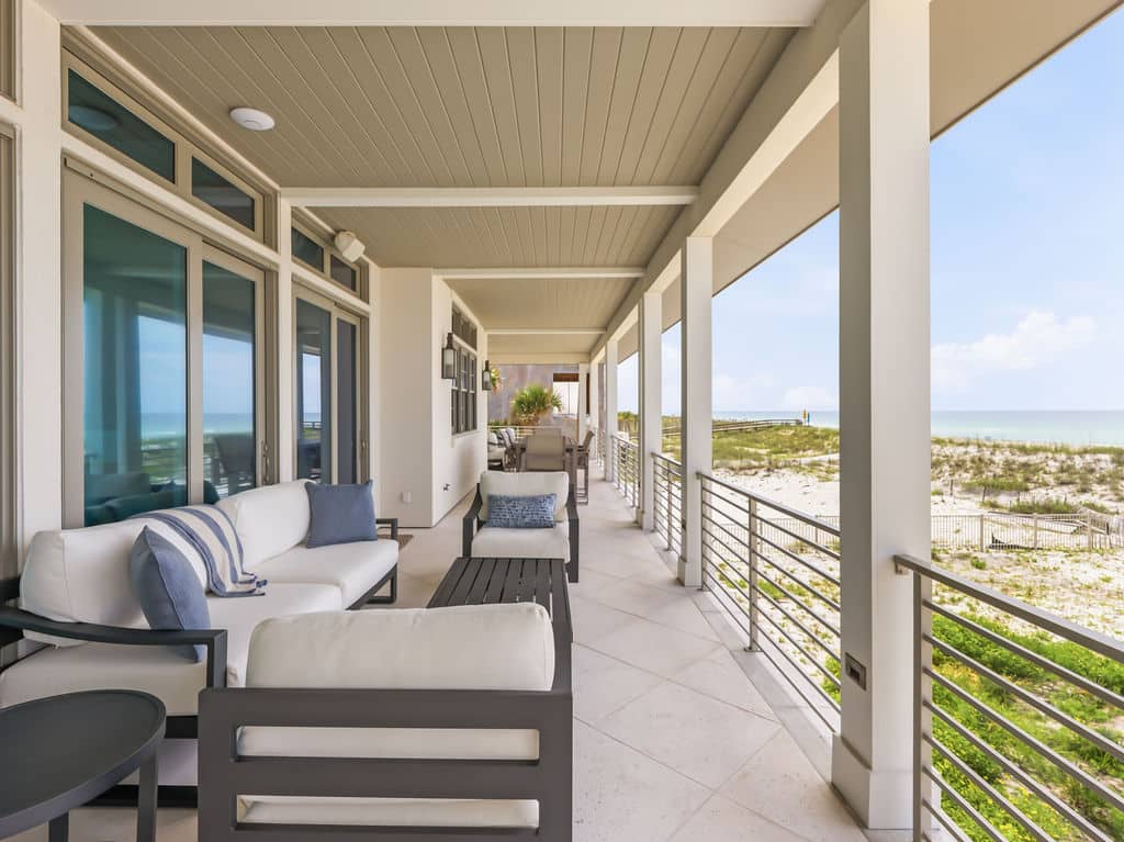 Covered coastal balcony featuring modern outdoor seating, tiled flooring, wood-paneled ceiling, and large glass sliding doors that face the ocean.