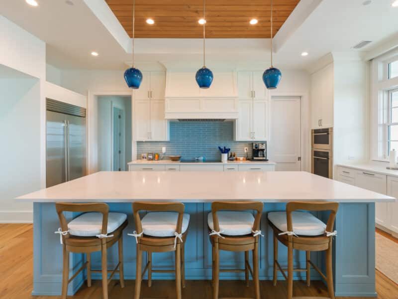 A large kitchen island with light blue cabinetry and four wooden barstools, set against a background of white cabinets, a blue subway tile backsplash, and three hand-blown blue glass pendant lights hanging from a wood-paneled ceiling recess.