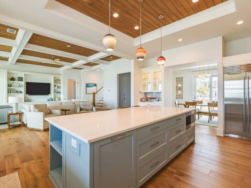 Open-concept luxury kitchen featuring a large light-blue island with a white quartz countertop, hand-blown colorful glass pendant lights, natural wood-paneled ceilings, and a view into the adjoining living room and dining area.