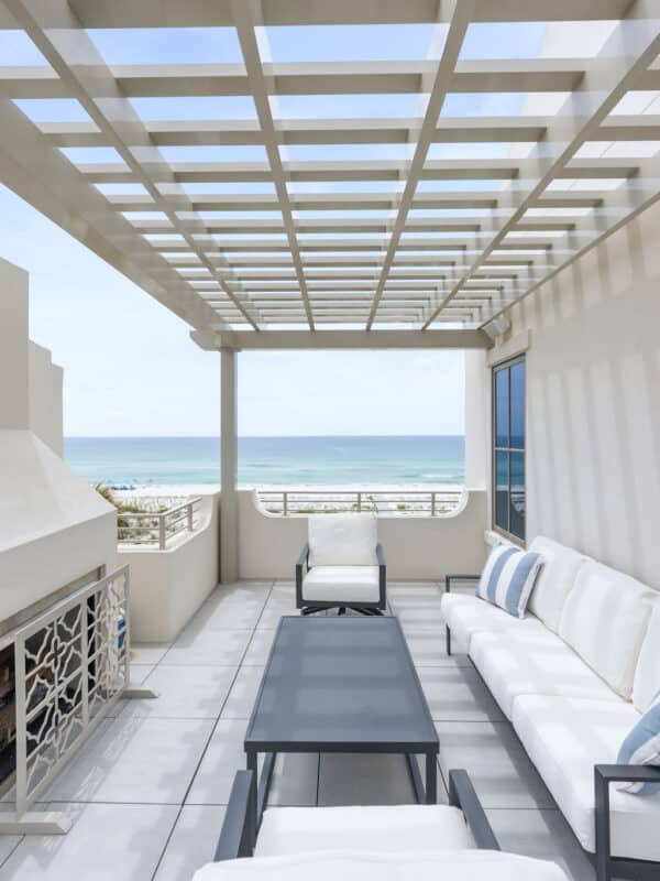 A modern beachfront rooftop patio featuring a white couch, a black coffee table, two white armchairs, an outdoor fireplace, and a beige wooden pergola overhead overlooking the ocean.