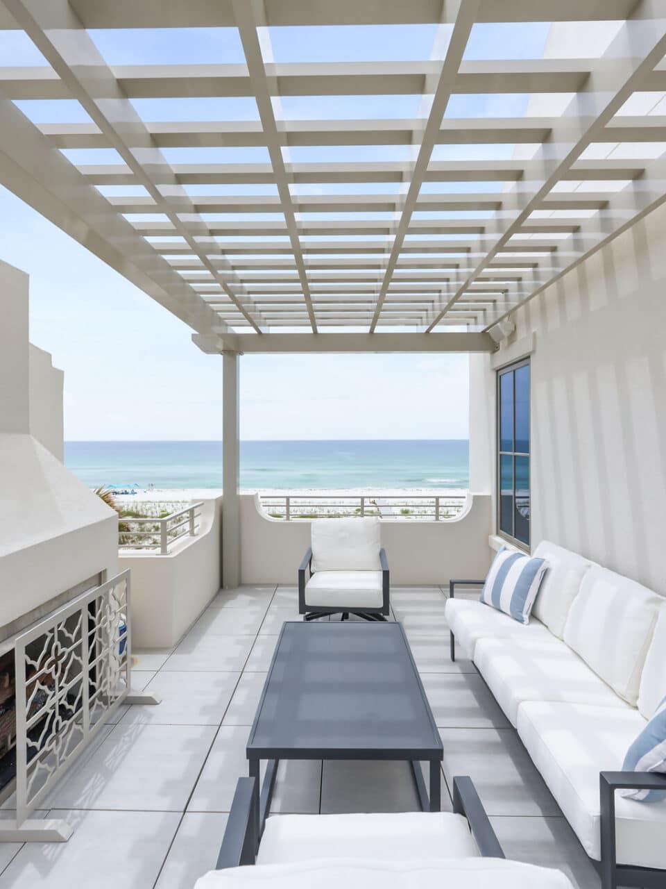 A modern beachfront rooftop patio featuring a white couch, a black coffee table, two white armchairs, an outdoor fireplace, and a beige wooden pergola overhead overlooking the ocean.