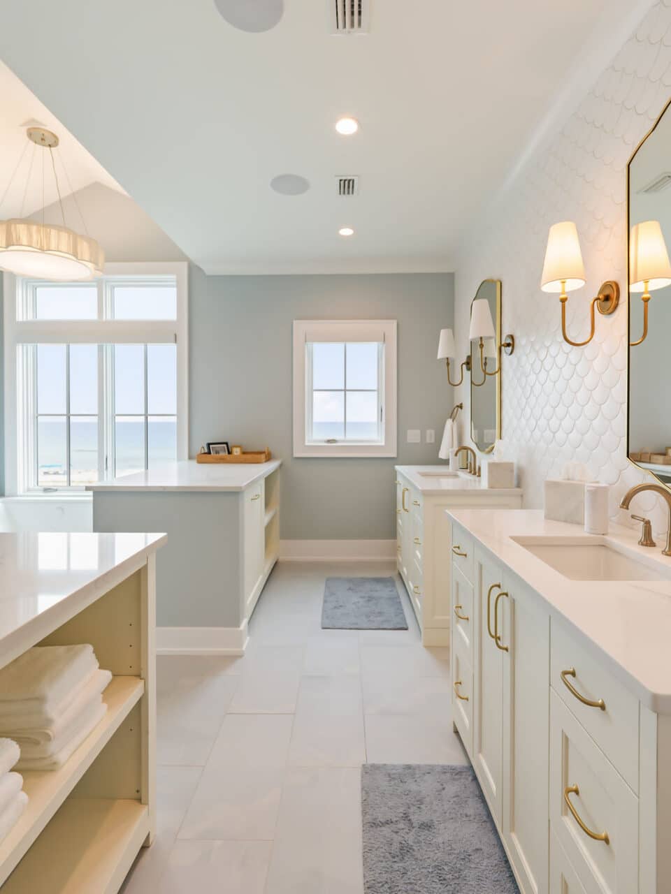 Bright coastal master bathroom with two cream vanities, gold fixtures, white scalloped tile backsplash, and a large window overlooking the ocean.