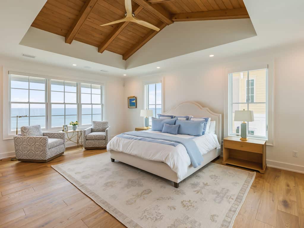 Bright coastal bedroom with a king bed, light blue accent pillows, a vaulted wood ceiling with a wooden fan, and two armchairs overlooking the ocean through large windows.