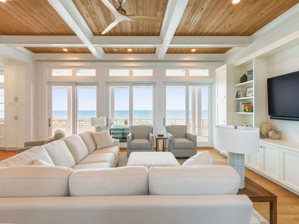 A bright, coastal-style living room featuring a large white sectional sofa, a wood-paneled coffered ceiling, and large glass doors with an ocean view.