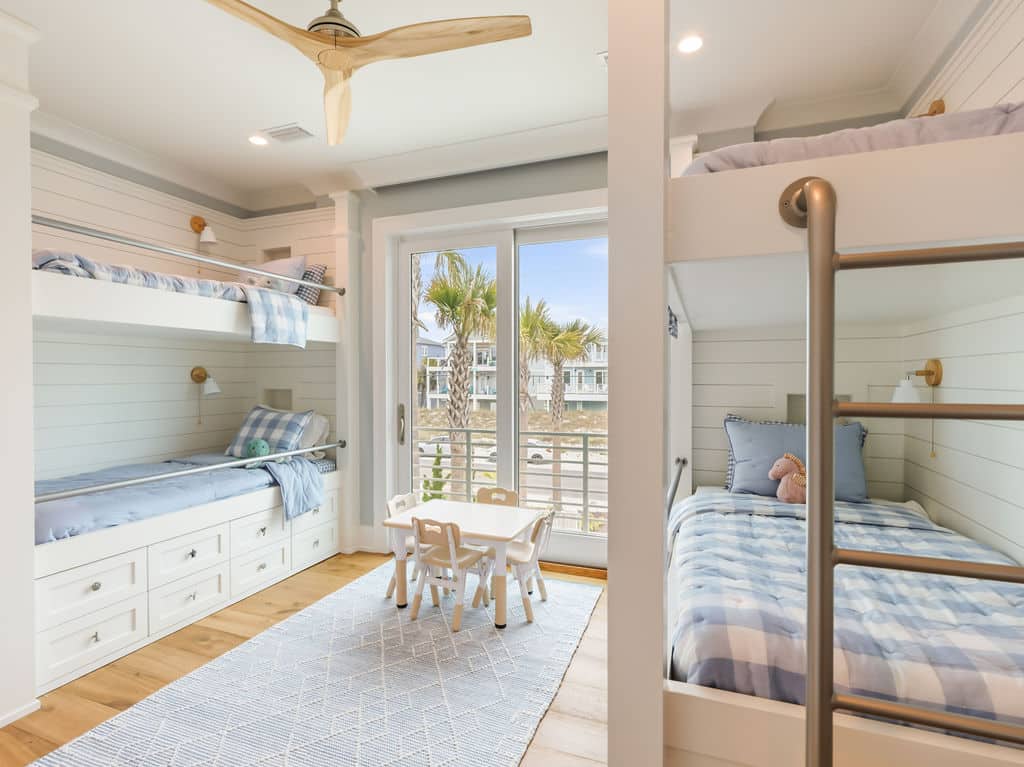 Modern coastal bunk bed room featuring built-in white shiplap bunk beds with blue plaid bedding, a small children's table and chairs, a wooden ceiling fan, and large sliding glass doors that open up to an ocean view.