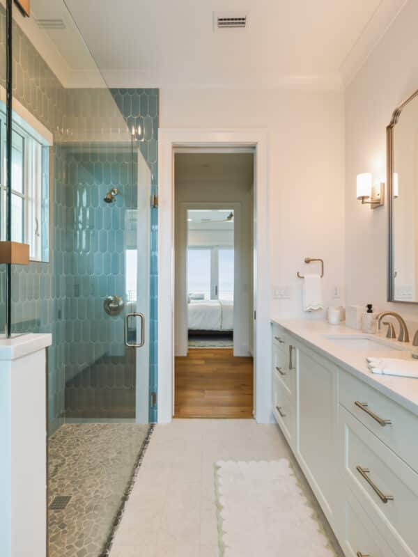 Modern primary bathroom featuring a glass walk-in shower with blue scalloped tile, a white vanity with gold hardware, and a doorway looking into a bedroom with an ocean view.