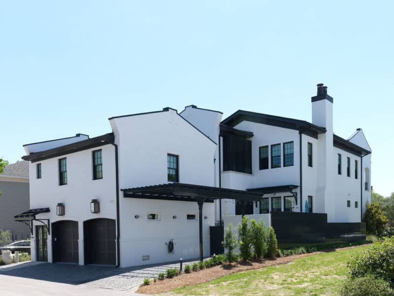 Side profile and rear exterior of a large white brick custom home featuring a two-car garage with dark arched doors, black window frames, a matching black pergola, and minimal landscaping.