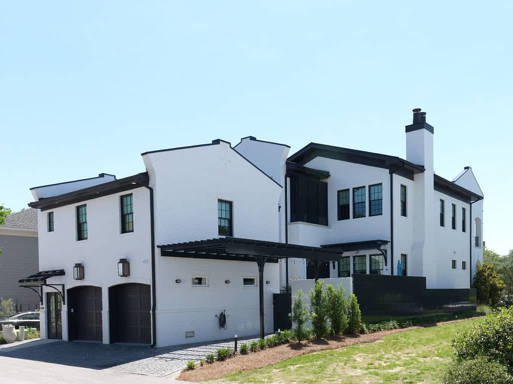 Side profile and rear exterior of a large white brick custom home featuring a two-car garage with dark arched doors, black window frames, a matching black pergola, and minimal landscaping.
